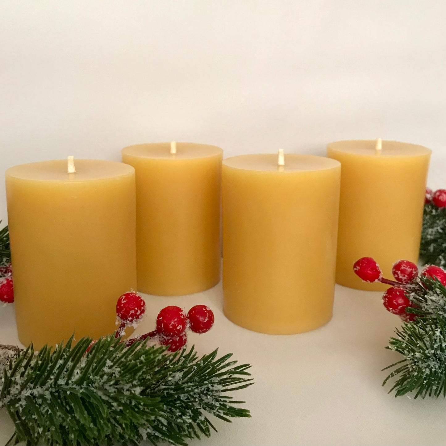 Four yellow beeswax candles with red berries and greenery on a white background