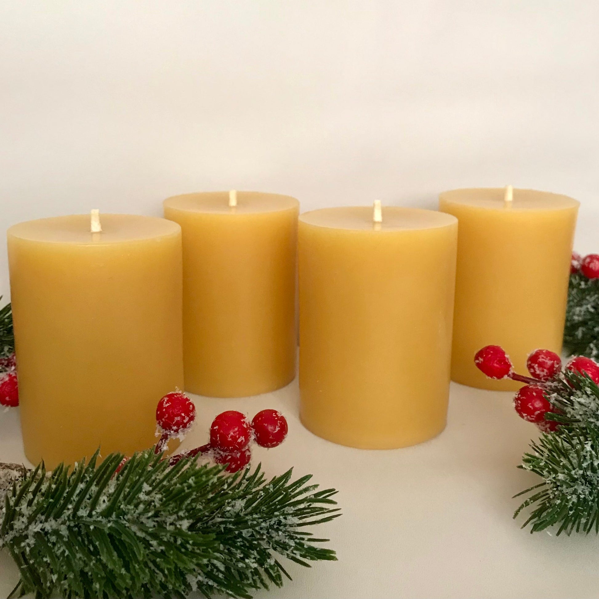 Four yellow beeswax candles with red berries and greenery on a white background
